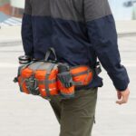 Person wearing a navy blue jacket and olive green pants carrying an orange and gray multifunctional waist bag with multiple compartments, including a water bottle holder, while walking outdoors, indicating the product’s utility for active lifestyles.