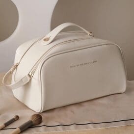 A cream-colored Double Zipper Toast Cosmetic Bag with a handle and the text "SELF OF WE MAKE CARE" embossed on the side, placed on a beige surface next to some makeup brushes.