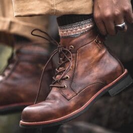 A person wearing tan pants and patterned socks is seen tying the laces of retro lace-up leather boots.