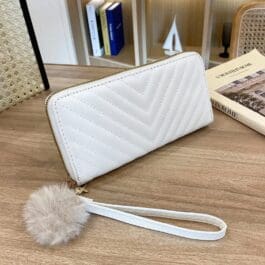 The wallet is displayed on a wooden surface, featuring a sleek white design with a chevron pattern. It appears to have a zip-around closure. Accompanying the wallet is a fluffy gray pom-pom attached to a white strap, adding an elegant yet playful touch to the accessory. The background includes soft-focus items such as books and another bag, suggesting the setting of a cozy interior space. This wallet is suitable for everyday use or as part of an ensemble for outings.