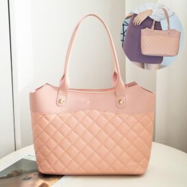 The handbag is a soft pink hue, featuring a quilted design with rhombus-shaped stitching. It has two sturdy handles attached with gold-tone hardware and a detachable crossbody strap (partially visible in the reflection of a mirror behind it). The bag rests on a white surface, and the blurred background provides no additional detail about the handbag itself.