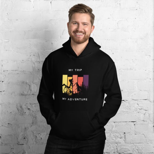 A smiling man with short hair wears a Versatile Unisex Hoodie, featuring a colorful graphic and the text My Trip, My Adventure, set against a pristine white brick wall.