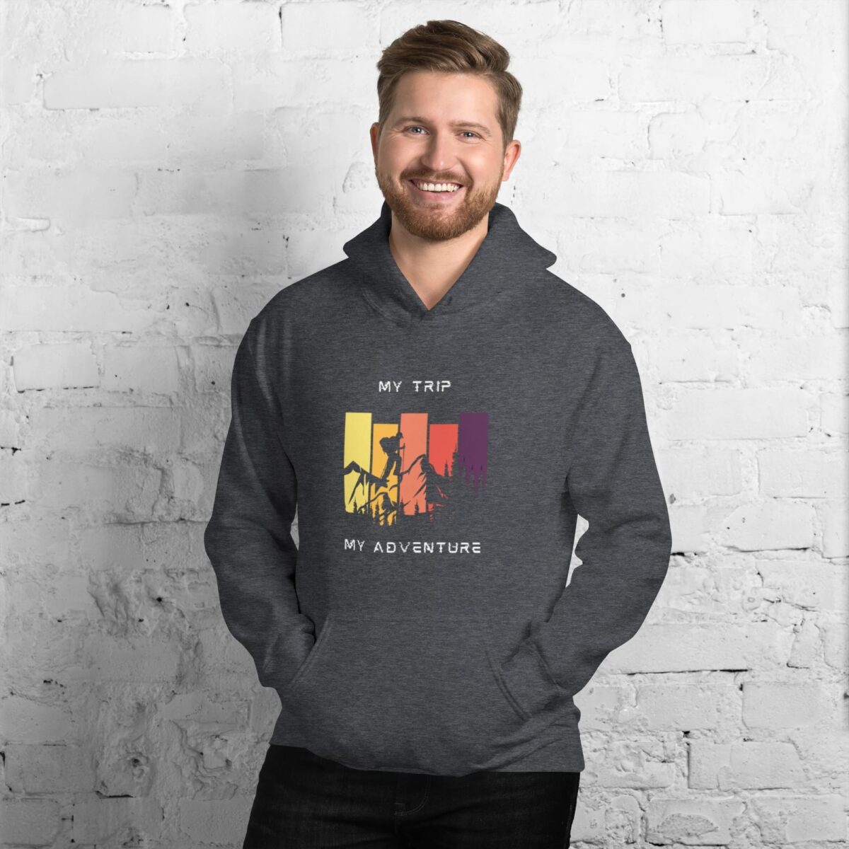 A bearded man smiles wearing the Versatile Unisex Hoodie, stylish for all genders, featuring a graphic and the text MY TRIP MY ADVENTURE, set against a white brick wall.