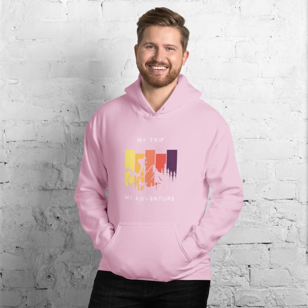 A smiling man with short brown hair and a beard wears the Versatile Unisex Hoodie, featuring colorful bars and the text My Trip, My Adventure, standing against a white brick wall.