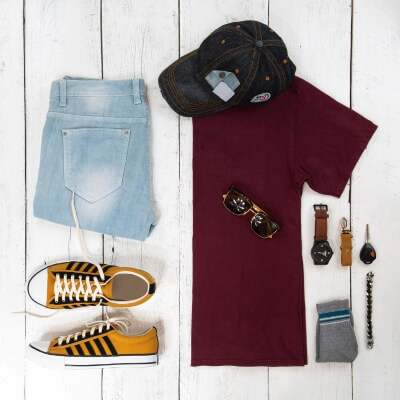 Men’s Clothing collection featuring a burgundy t-shirt, light blue jeans, yellow and black sneakers, a dark cap, sunglasses, a brown leather watch, gray socks, and a bracelet on a white wooden background