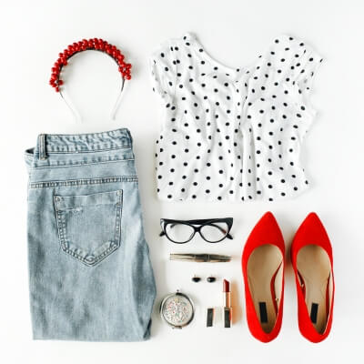 Fashionable Women’s Clothing flat lay with polka dot top, blue jeans, red high heels, accessories including sunglasses, watch, and makeup on a white background.