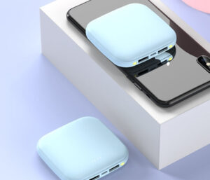 A modern wireless charging pad with a light blue top is placed on a white surface, charging a black smartphone that lies partially on the pad. Another identical wireless charging pad is in the foreground, suggesting the convenience of multiple-device charging capability. The image has a soft pastel background which gives it a clean and minimalist aesthetic.